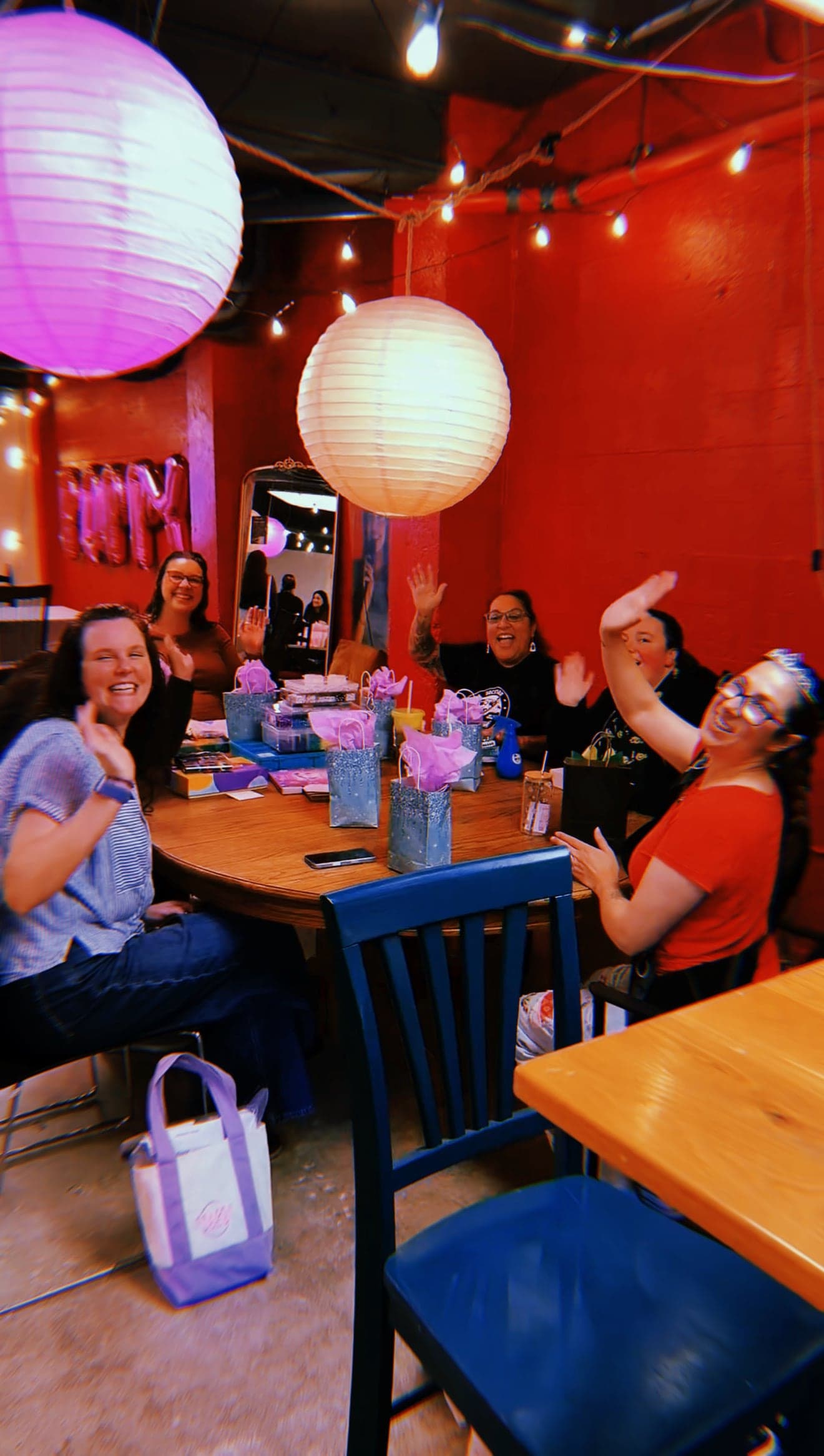 A party group waving from a table set with gift bags under paper lanterns